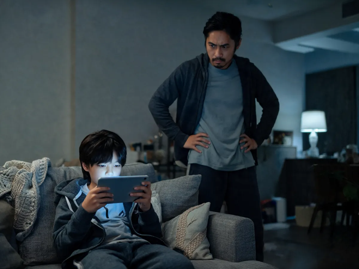 A child absorbed in a tablet on the couch while a parent looks on, worn out